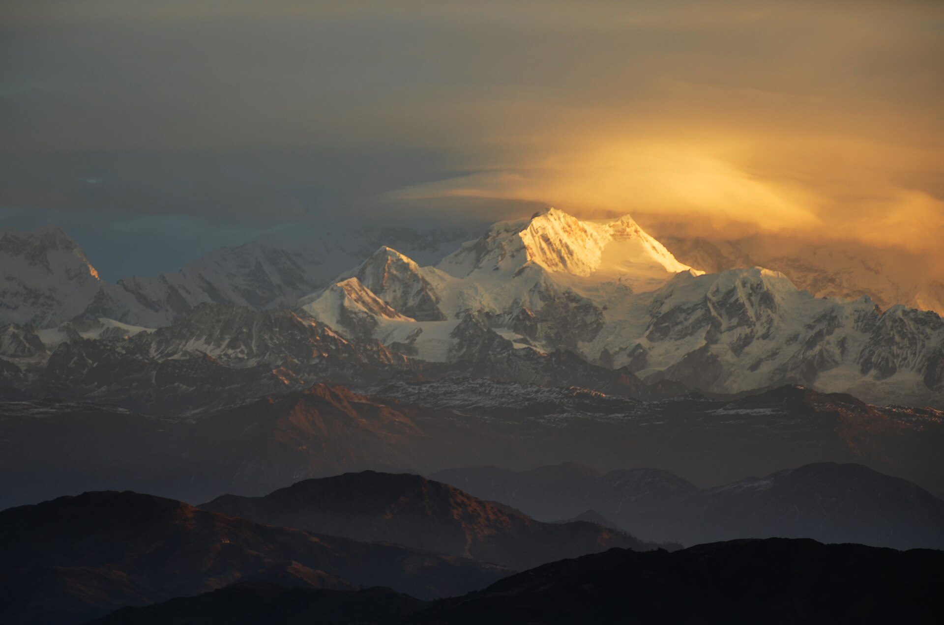 Kangchenjunga massif illuminated by warm golden sunrise light above layered clouds and dark Himalayan ridges