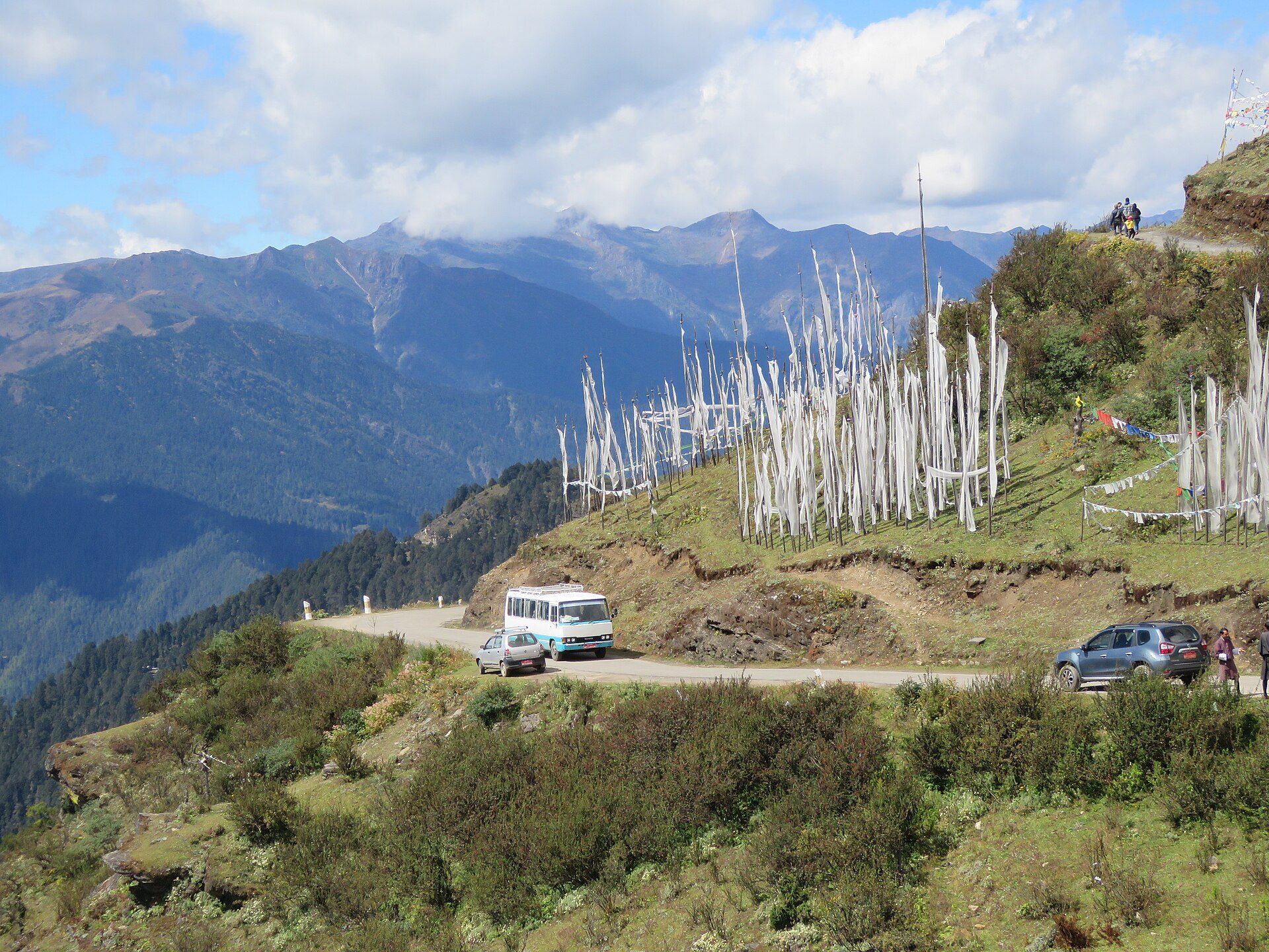 Clusters of tall white Himalayan prayer flags lining a winding high mountain pass road against distant blue ridges