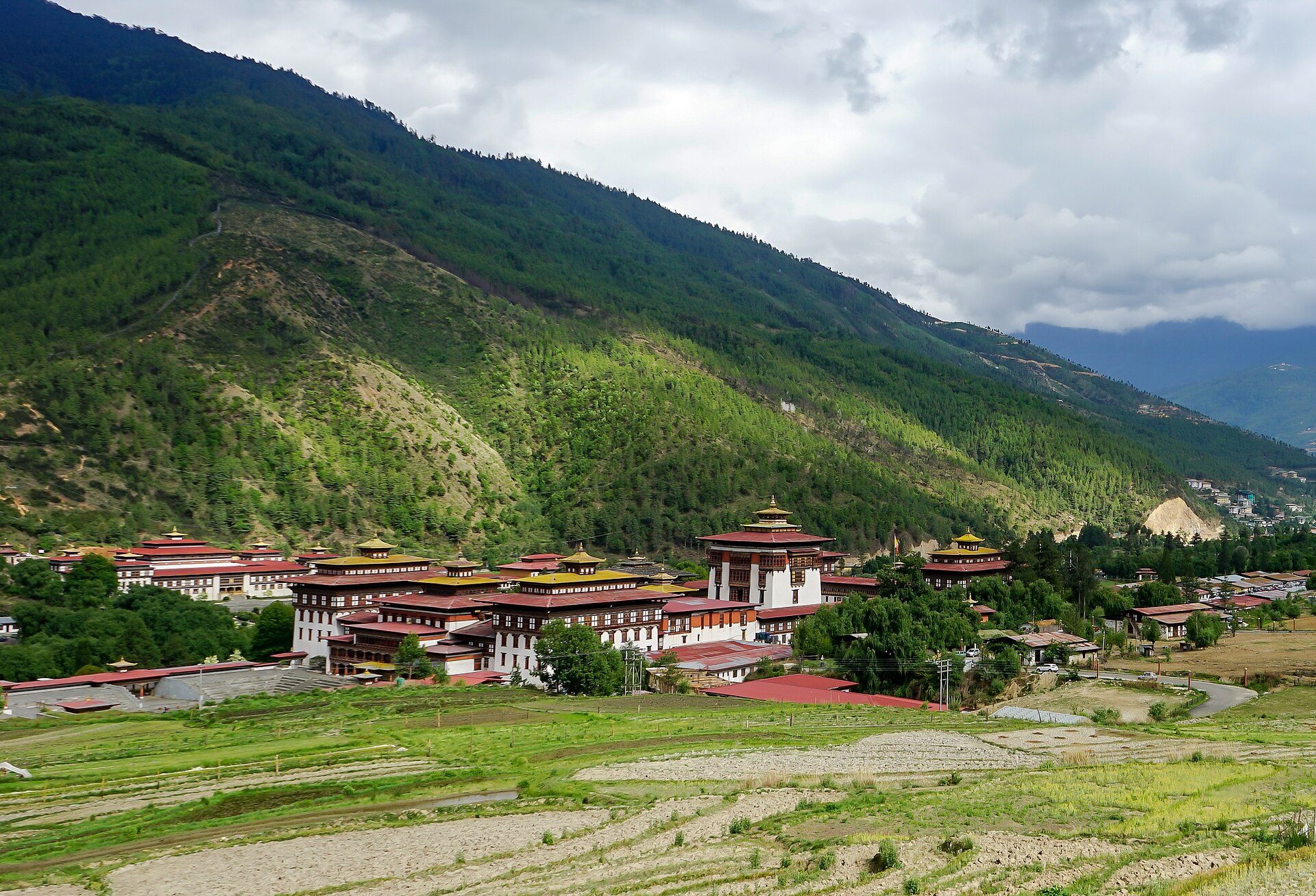 Whitewashed walls and traditional tiered roofs of Tashichho Dzong set beside terraced fields and forested mountains in Thimphu, Bhutan